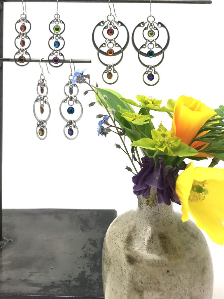 A rainbow of flowers in a small vase in Wraptillion's studio, with jewelry from the Industrial Glass Rainbows collection: poppies, columbine, euphorbia, hosta, forget-me-nots.