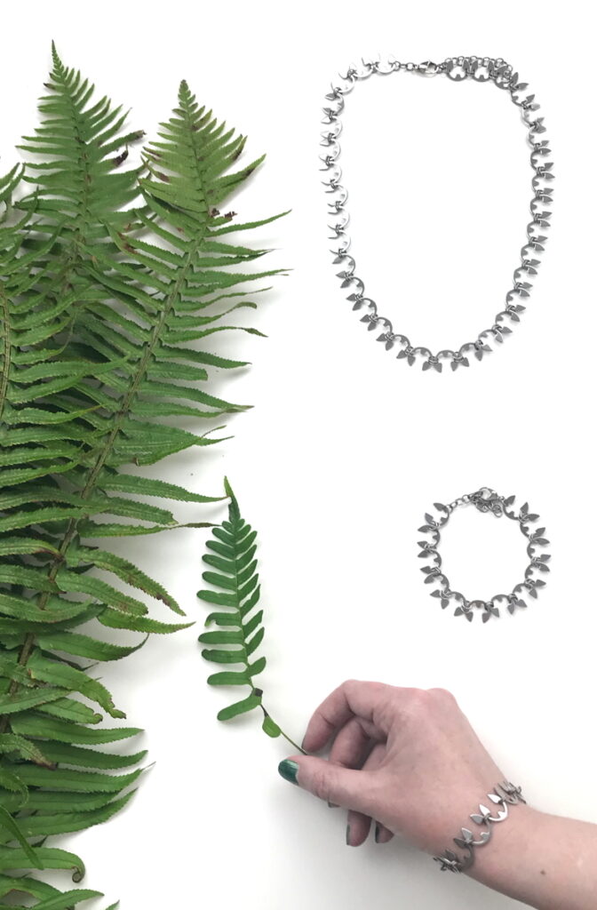 Cut sword fern fronds, shown in a flat lay with the Fern Necklace and Fern Bracelet. A hand wearing the Fern Bracelet holds another fern frond.