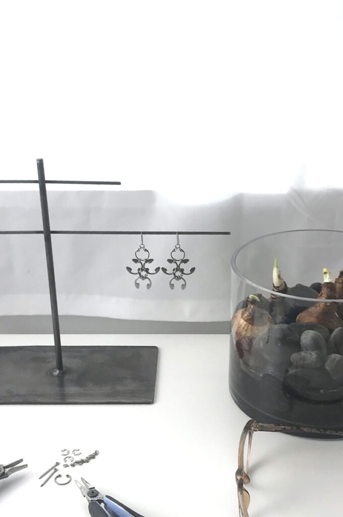Wraptillion's desk, with reading glasses, paperwhite narcissus bulbs sprouting amid rocks in a shallow modern glass vase, pliers, a jewelry project in progress, and the Garland Earrings.