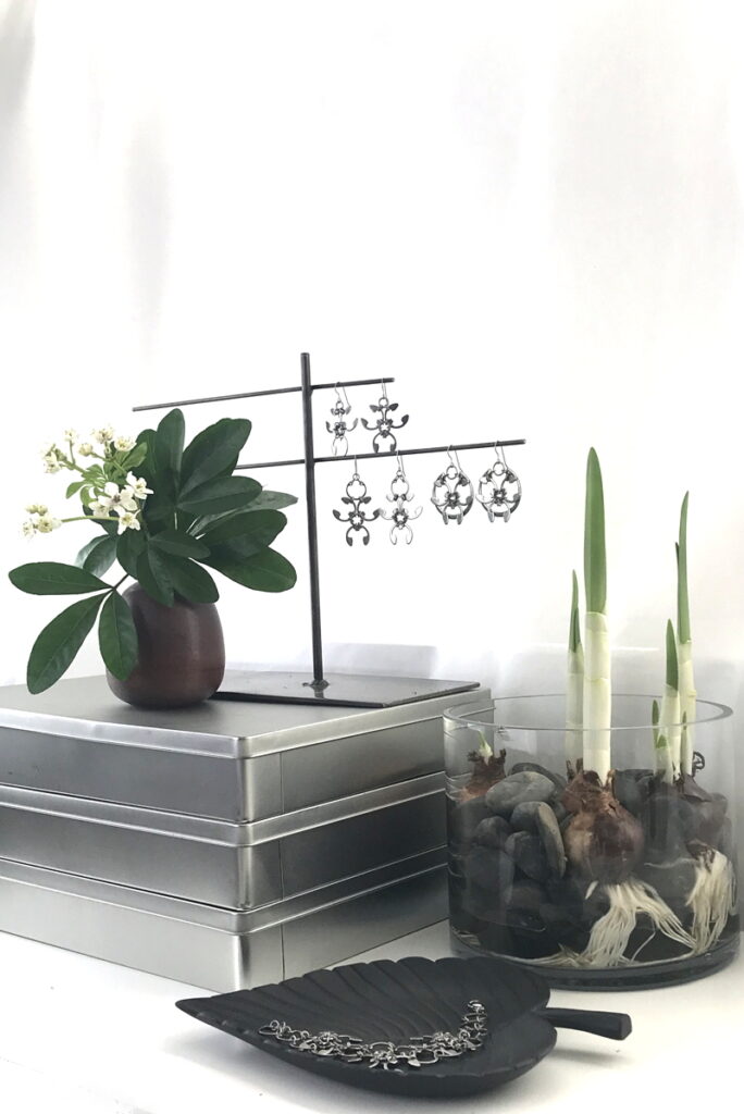 Choisya ternata flowers and leaves in a small modern bud vase on a stack of metal boxes, next to Wraptillion's Trellis Earrings, Garland Earrings, and Rose Window Earrings. Paperwhite narcissus are growing amid rocks in a glass vase. A black leaf dish holds the Garland Bracelet. A sneak peek into Wraptillion's studio.
