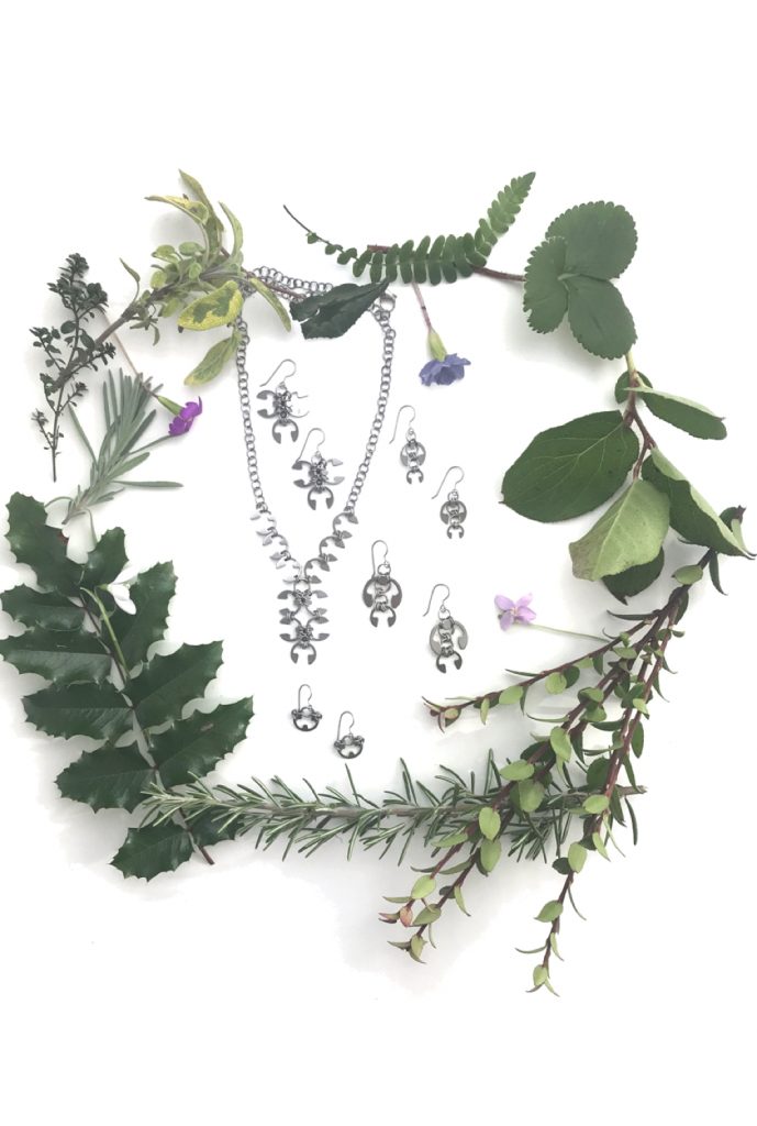 Botanical-inspired jewelry from Wraptillion's Mechanical Garden collection, surrounded by branches from mahonia, salal, rosemary, and other evergreen plants, with snowdrop, primrose, and violet spring flowers.