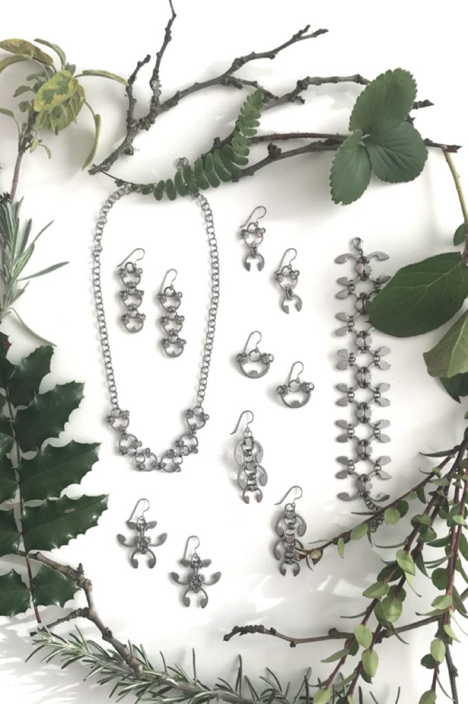 Closeup of botanical-inspired jewelry from Wraptillion's Mechanical Garden collection, surrounded by branches from plum, mahonia, salal, rosemary, and other evergreen plants.in winter.