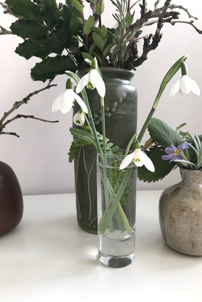 cut snowdrops in a small glass vase with a fern frond, shown in front of other small winter floral arrangements