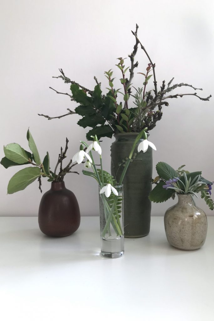 cut snowdrops in a small glass vase with a fern frond, shown in front of 3 other small winter floral arrangements