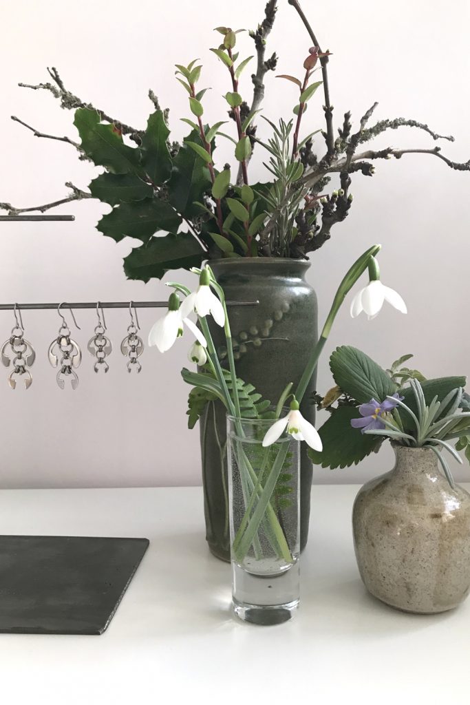 cut snowdrops in a small glass vase with a fern frond, shown in front of other small winter leaf, branch, and flower arrangements and Wraptillion's Hops Earrings and Short Fuchsia Earrings