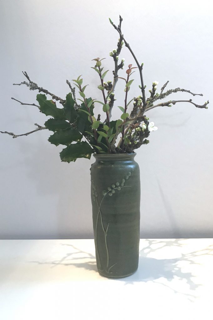 Photo of a small casual winter floral arrangement in a green ceramic vase, with mahonia, evergreen huckleberry, and plum branches breaking into flower.