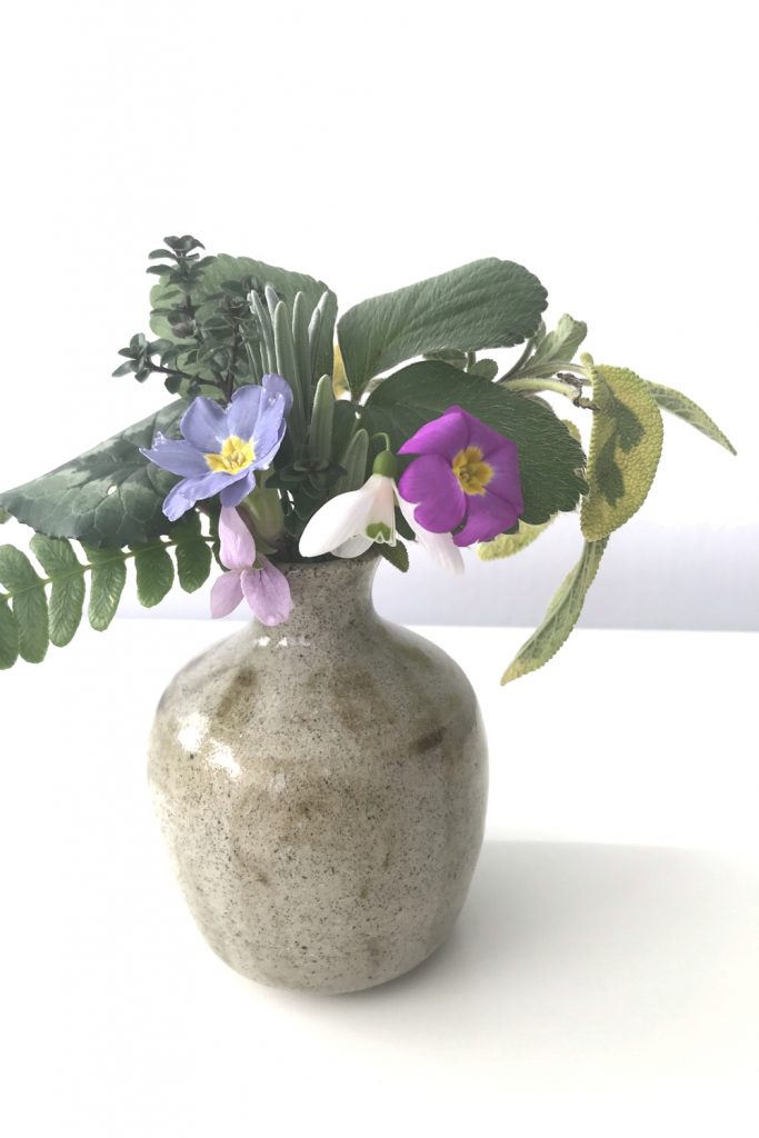 Wraptillion's studio flowers: a small everyday winter flower arrangement with strawberry, thyme, sage, lavender, fern, primrose, snowdrop, & violet, in a small beige handmade ceramic vase.