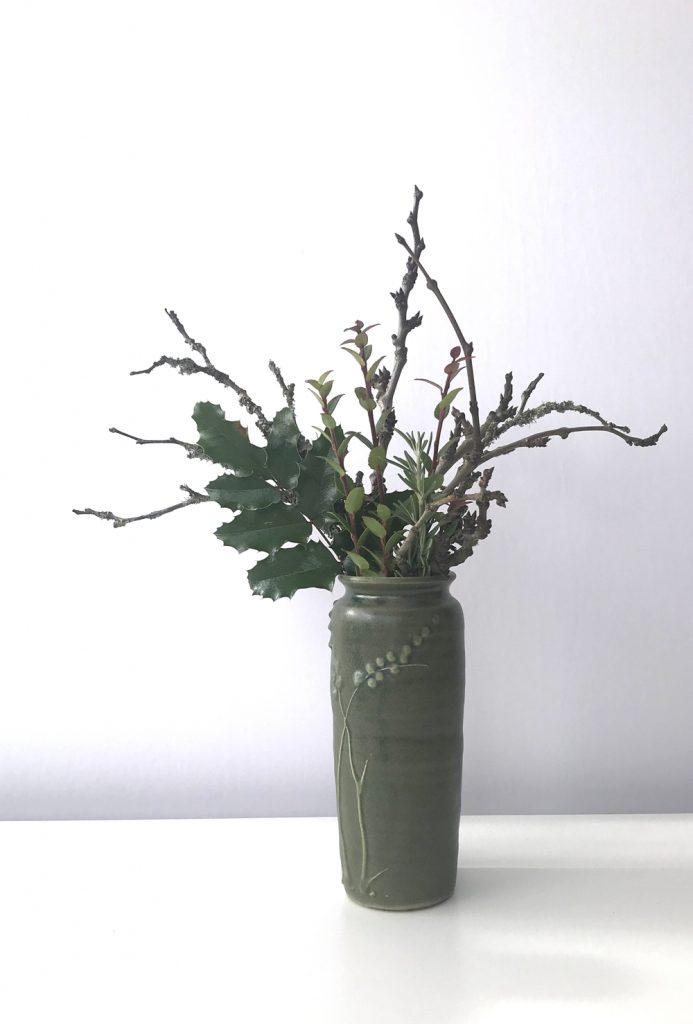 Waptillion's studio flowers: a small everyday winter flower arrangement, featuring bare plum branches and evergreen huckleberry, mahonia, and rosemary leaves, in a handmade green ceramic vase.