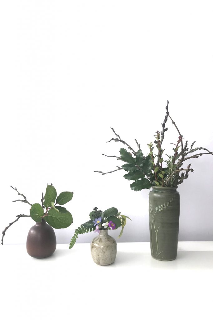 Three small everyday winter flower arrangements, featuring bare plum branches, evergreen leaves such as salal and mahonia, and a few early spring flowers like primrose, snowdrop, and violet, all in ceramic vases.