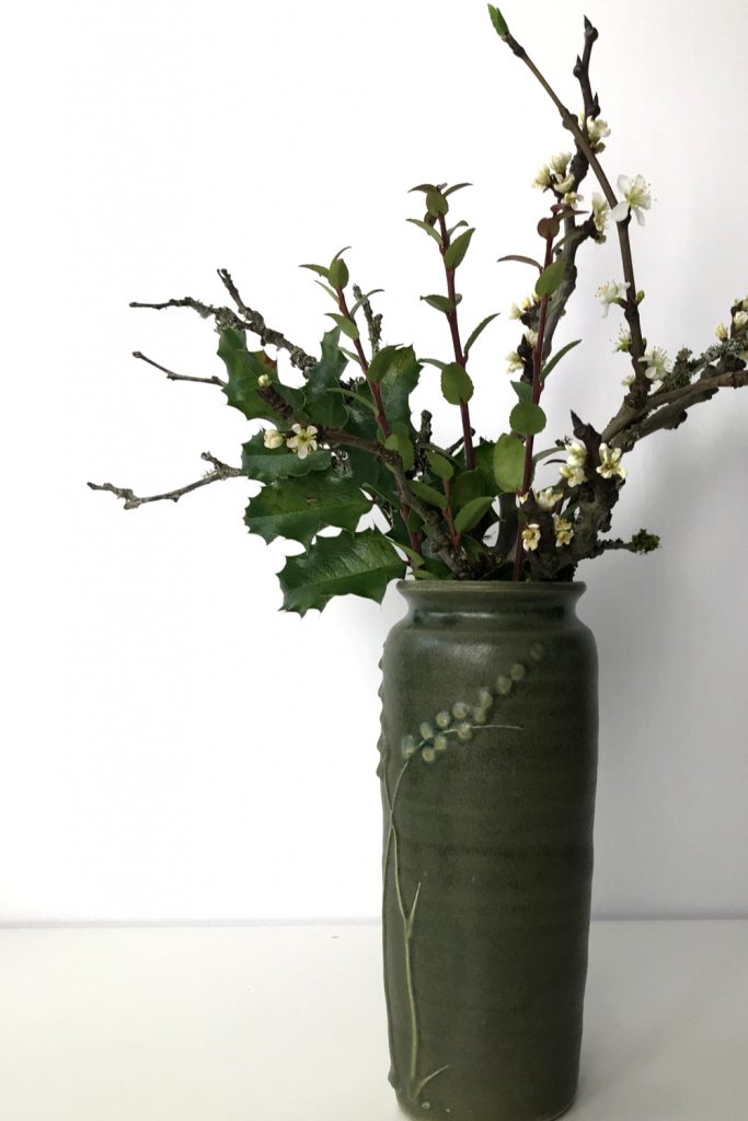 A small winter arrangement of plum branches in bloom with mahonia and huckleberry leaves in a green ceramic vase.