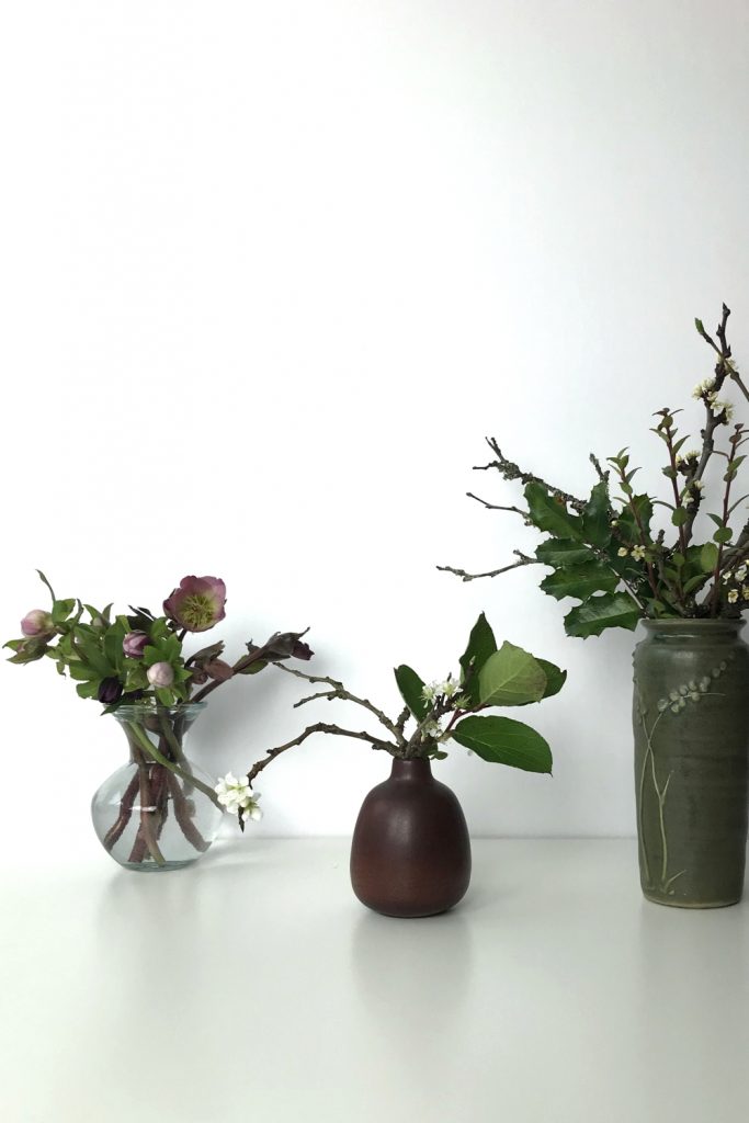 Three small casual winter flower arrangements: pink hellebores in a glass vase, flowering plum branches and salal leaves in a brown bud vase, plum branches, mahonia, and huckleberry leaves in a green vase.