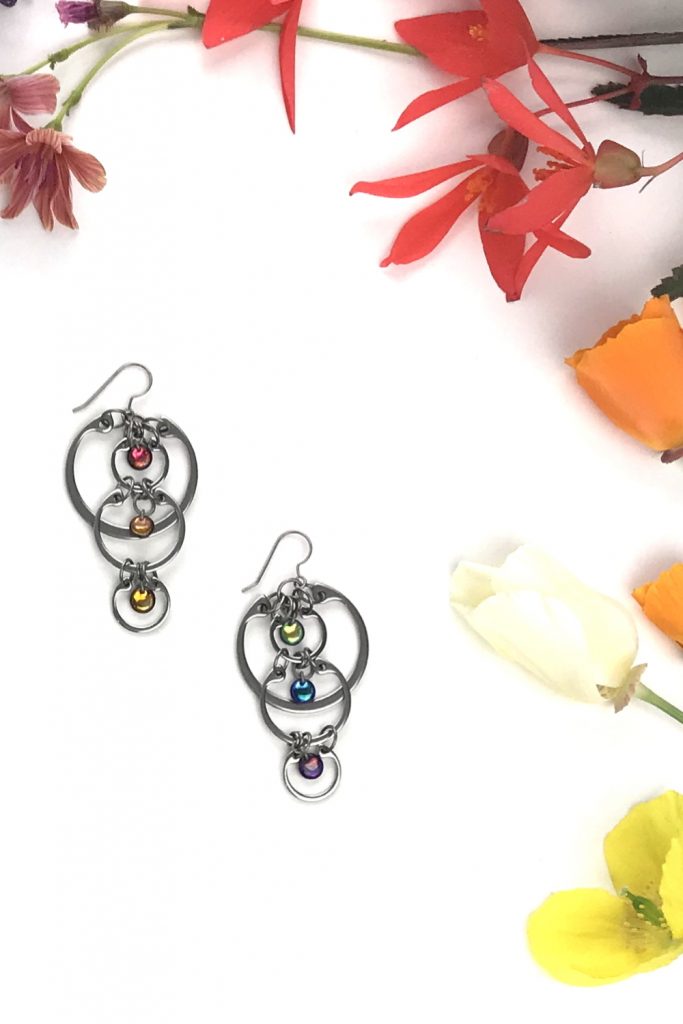 Wraptillion's modern Cascading Rainbows Earrings in a flatlay next to yellow Welsh poppies, cream and orange California poppies, red begonias, and coral lewisia flowers.