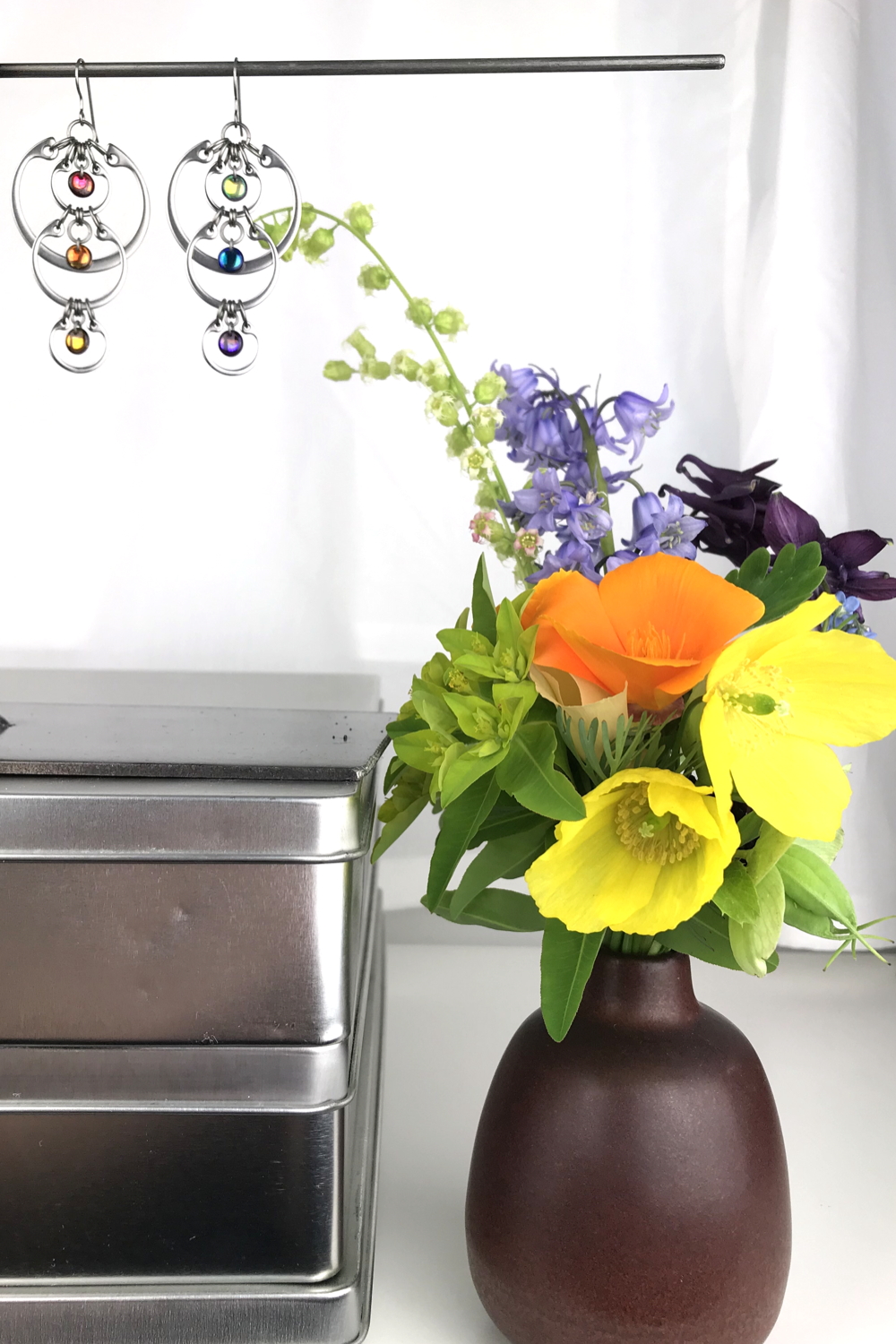 A jewelry stand with Wraptillion's modern Cascading Rainbow Earrings sits on a stack of metal boxes, next to a ceramic bud vase with bluebells, poppies, fringe flowers, and euphorbia in a studio.