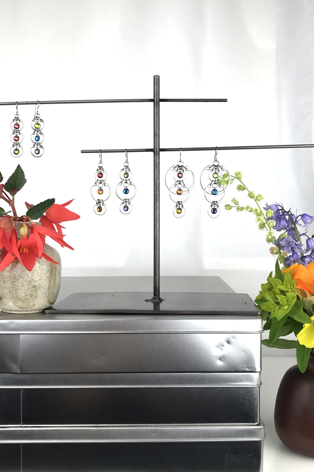 A ceramic bud vase of red begonia flowers sits on a stack of metal boxes, next to a jewelry stand with Wraptillion's modern Tripled Rainbow Earrings, Alternating Rainbow Earrings, and Cascading Rainbow Earrings. Another ceramic bud vase with bluebells, poppies, fringe flowers, and euphorbia sits nearby in a studio.