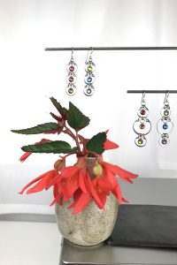 A ceramic bud vase of red begonia flowers sits on a metal box, next to a jewelry stand with Wraptillion's modern Tripled Rainbow Earrings and Alternating Rainbow Earrings, in a studio.
