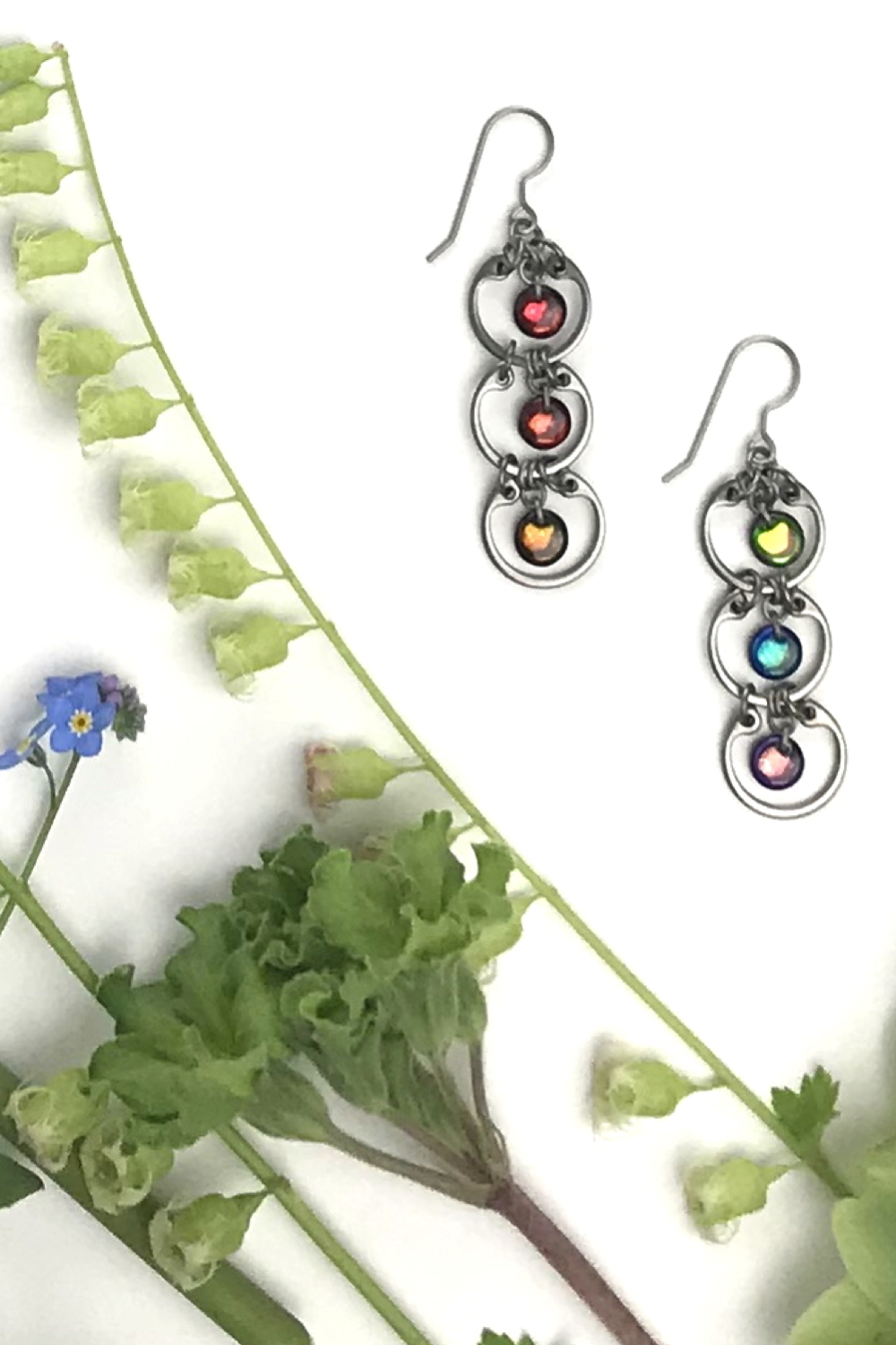 Wraptillion's modern Tripled Rainbow Earrings in a flatlay next to green fringe flowers and primroses and blue forget-me-nots.