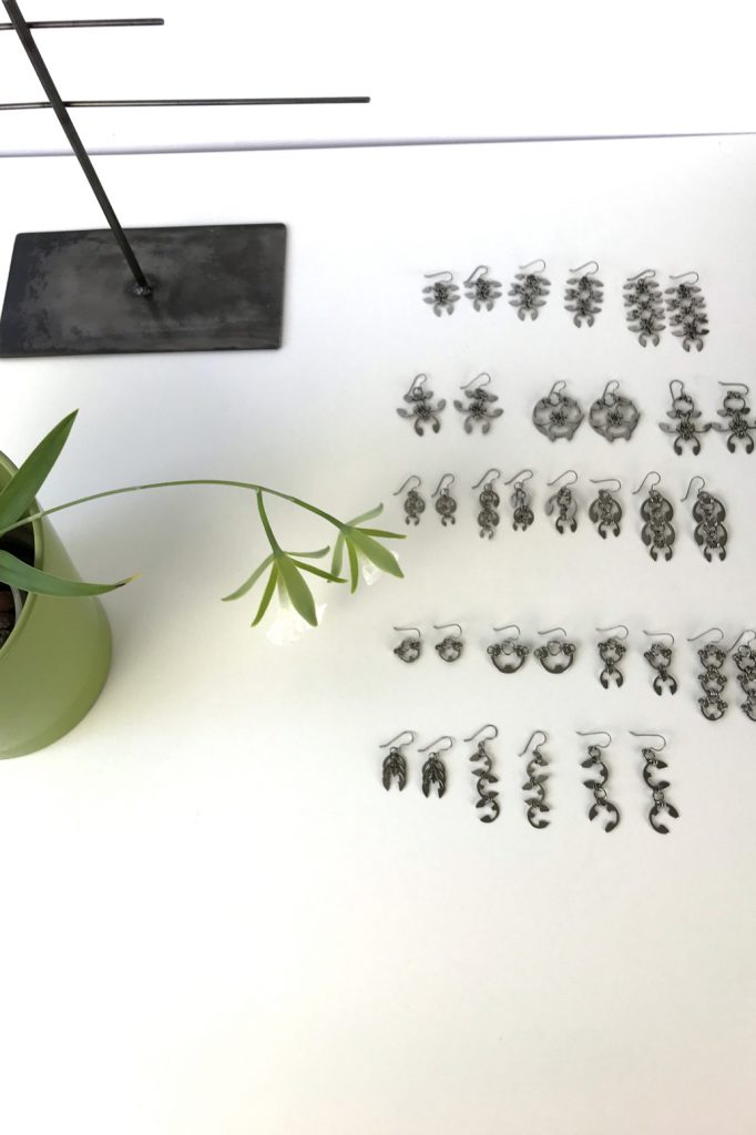 Orchid Encyclia mariae (formerly Euchile mariae) blooms on a desk in Wraptillion's studio next to a steel jewelry stand and earrings from Wraptillion's Mechanical Garden collection.