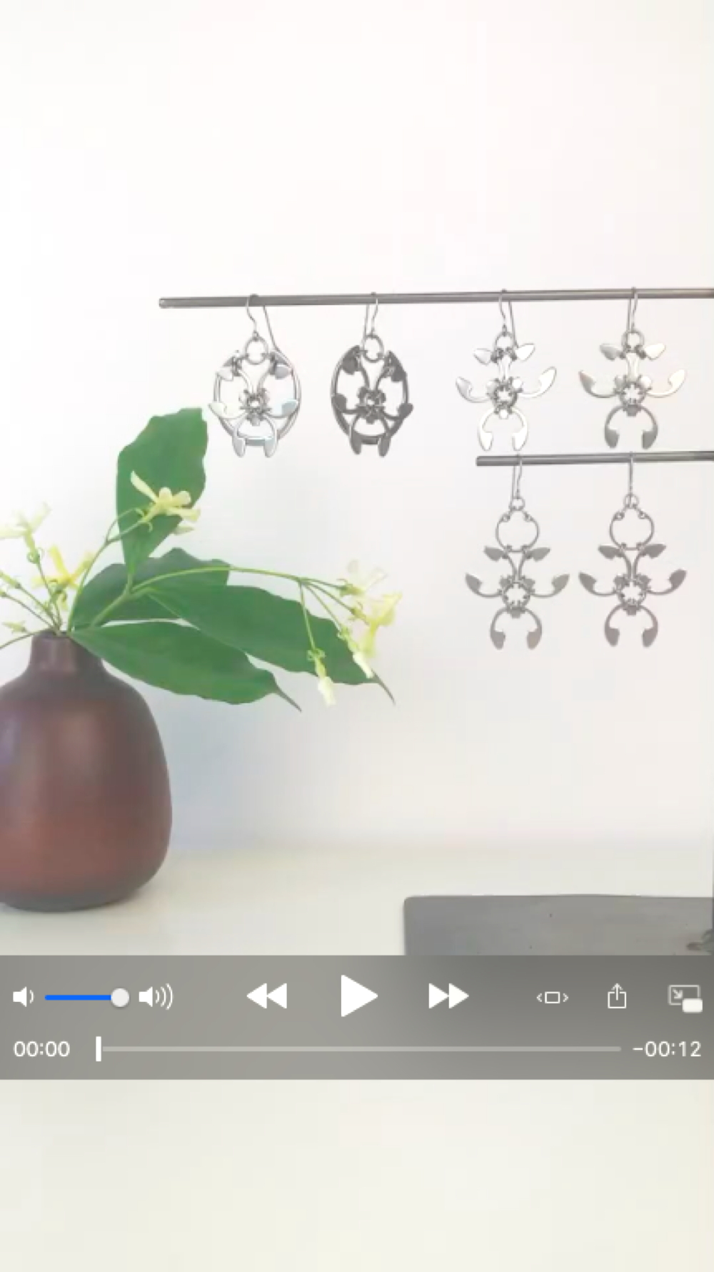 Cover screen for a short video featuring pale yellow star jasmine (Trachelospermum asiaticum) flowers and green leaves in a bud vase by Heath Ceramics on a desk in Wraptillion's studio workspace, with the modern botanical-inspired Rose Window Earrings, Trellis Earrings, and Garland Earrings.