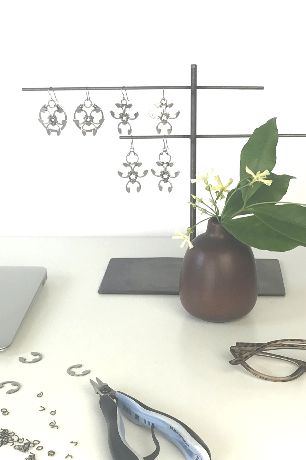 Pale yellow star jasmine (Trachelospermum asiaticum) flowers and green leaves in a bud vase by Heath Ceramics on a desk in Wraptillion's studio workspace, with the modern botanical-inspired Rose Window Earrings, Trellis Earrings, and Garland Earrings, reading glasses, pliers, and jewelry components..