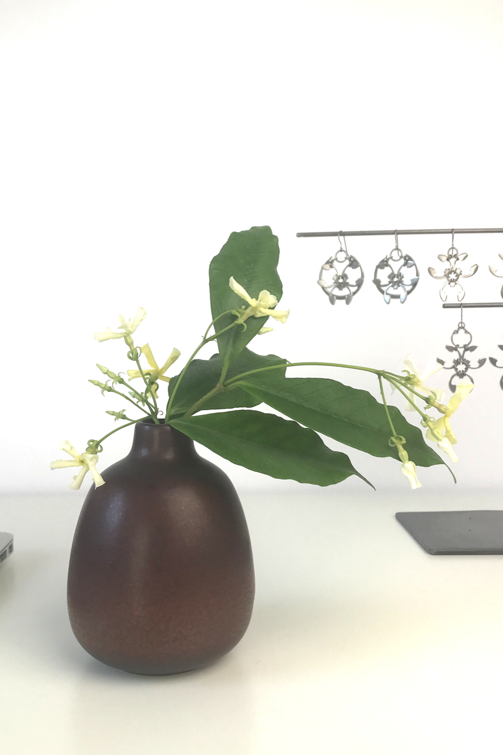 Pale yellow star jasmine (Trachelospermum asiaticum) flowers and green leaves in a bud vase by Heath Ceramics on a desk in Wraptillion's studio workspace, with the modern botanical-inspired Rose Window Earrings, Trellis Earrings, and Garland Earrings.