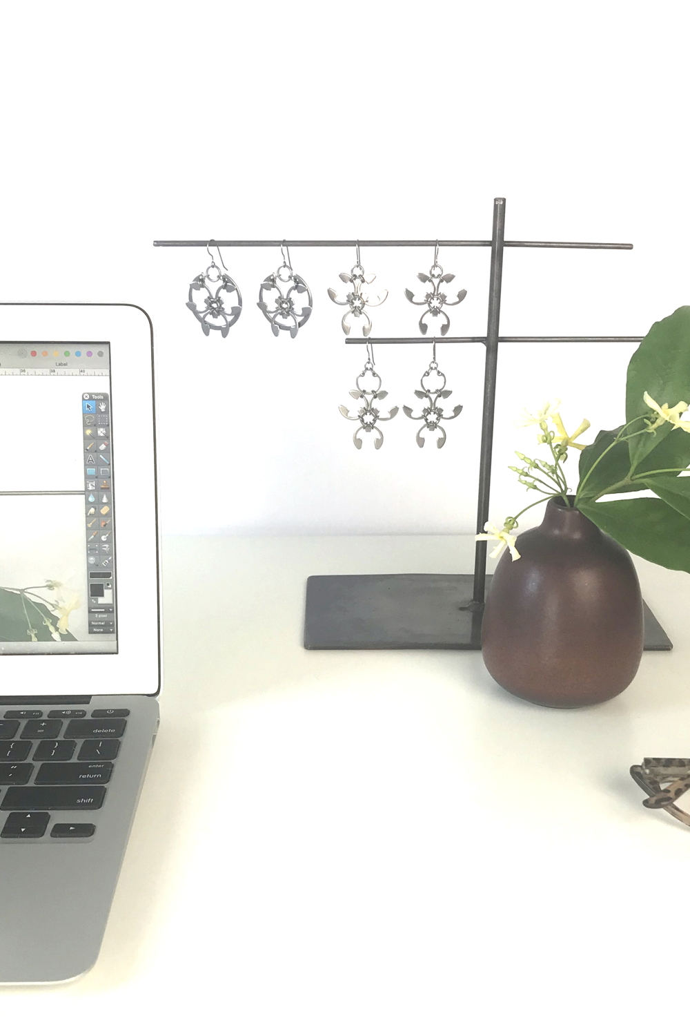 Pale yellow star jasmine (Trachelospermum asiaticum) flowers and green leaves in a bud vase by Heath Ceramics on a desk in Wraptillion's studio workspace, with the modern botanical-inspired Rose Window Earrings, Trellis Earrings, and Garland Earrings, reading glasses, and an open laptop..