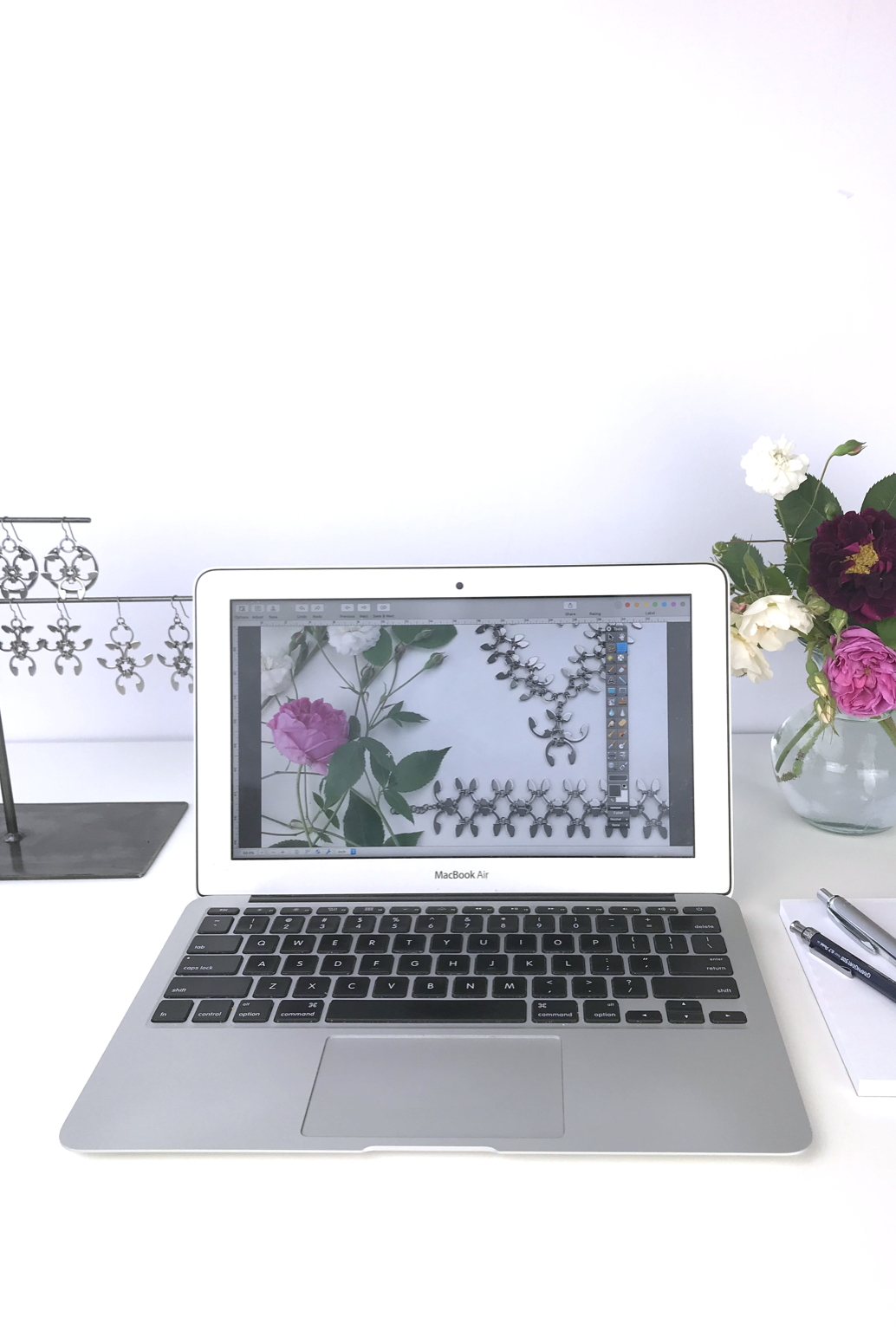 Open laptop next to a small arrangement of old roses (Rosa alba, Rosa gallica officinalis, moss rose 'Capitaine John Ingram', & 'Felicite et Perpetue') with earrings from the Mechanical Garden collection in Wraptillion's studio.