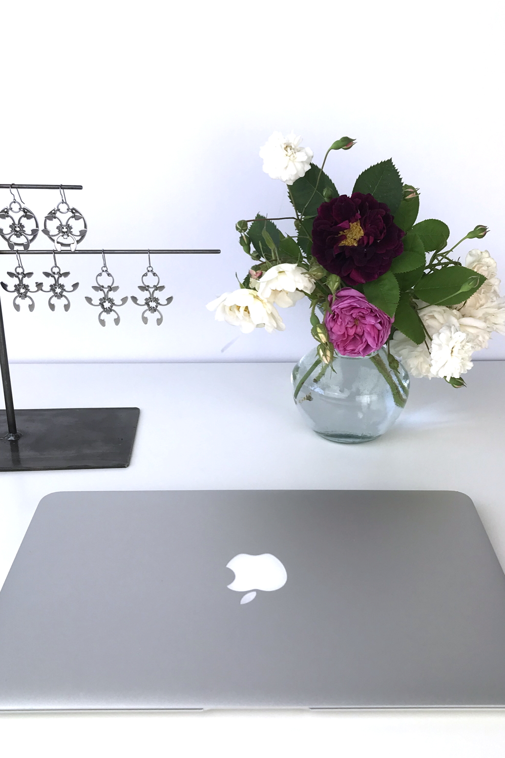 Closed laptop next to a small arrangement of old roses (Rosa alba, Rosa gallica officinalis, moss rose 'Capitaine John Ingram', & 'Felicite et Perpetue') with earrings from the Mechanical Garden collection in Wraptillion's studio.