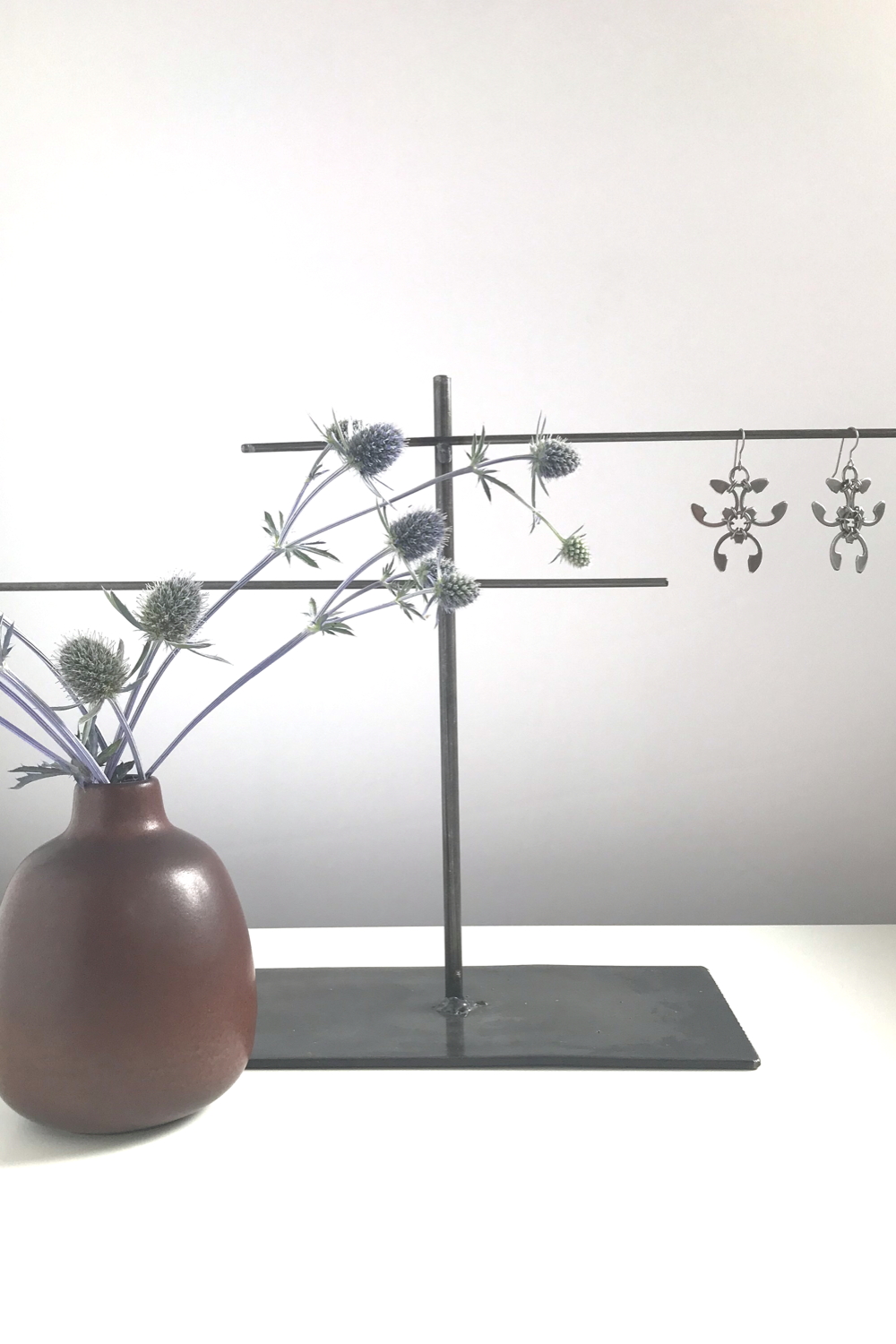 Sea holly (Eryngium) in a brown bud vase by Heath Ceramics on a desk next to a jewelry stand with Wraptillion's spiky floral Trellis Earrings.