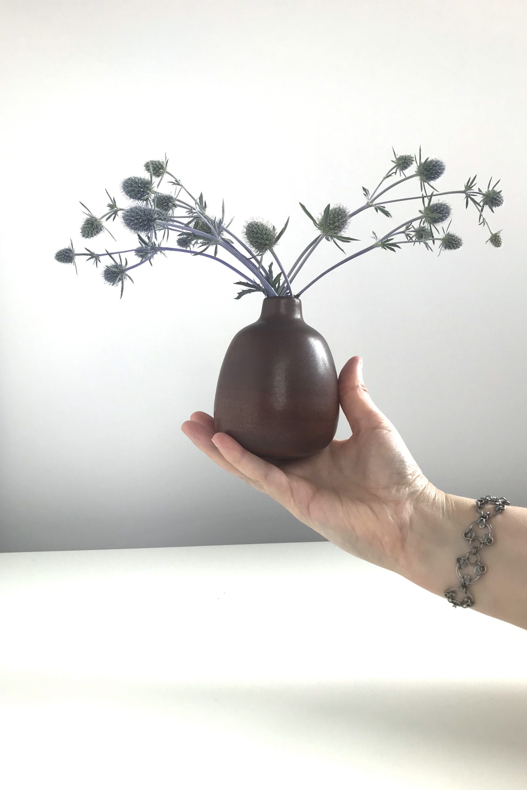 A hand holding a brown bud Heath Ceramics bud vase of sea holly (Eryngium), wearing Wraptillion's intricate modern chainmaille Scarab Bracelet..