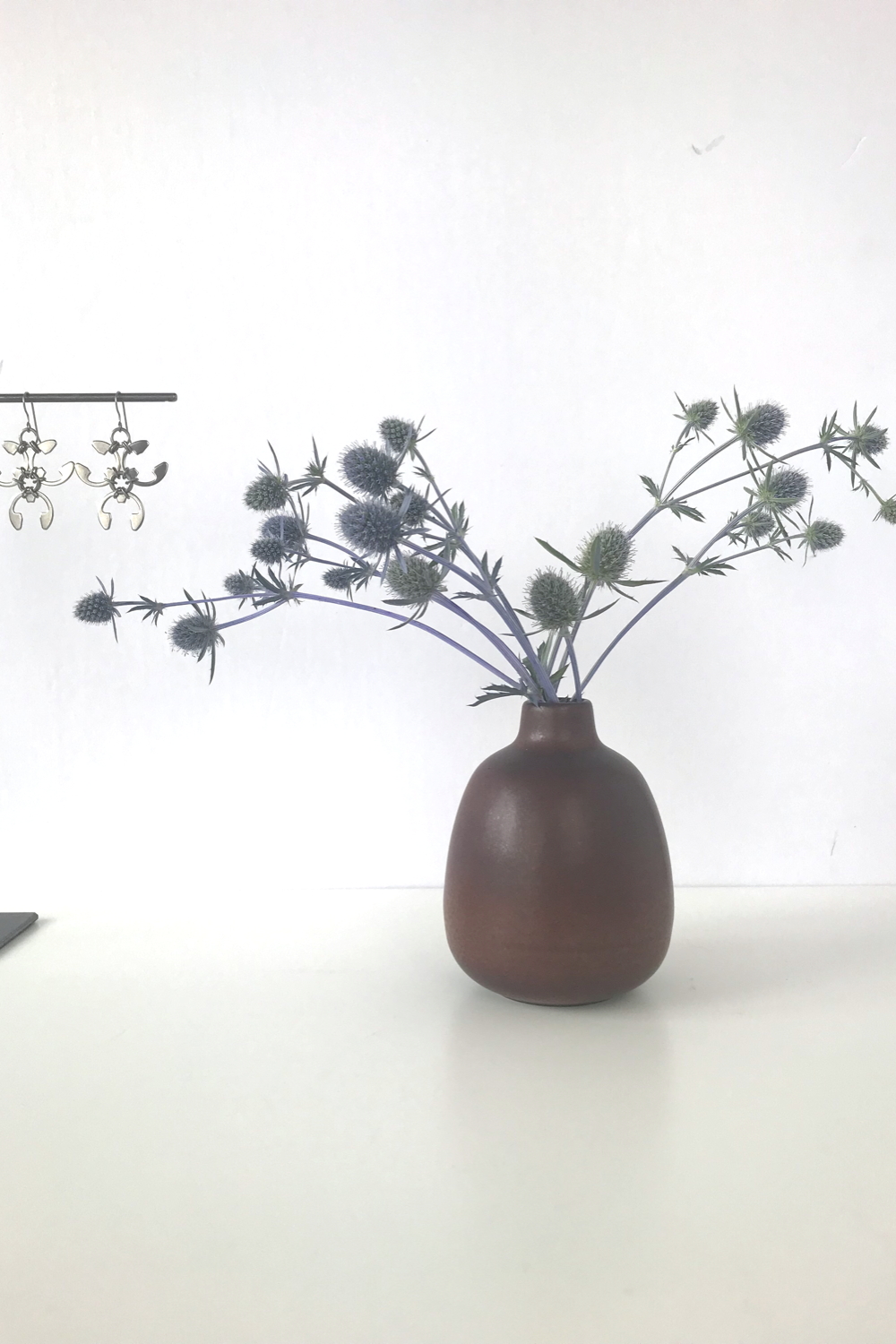 Sea holly (Eryngium) in a brown bud vase by Heath Ceramics on a desk next to a jewelry stand with Wraptillion's spiky floral Trellis Earrings.