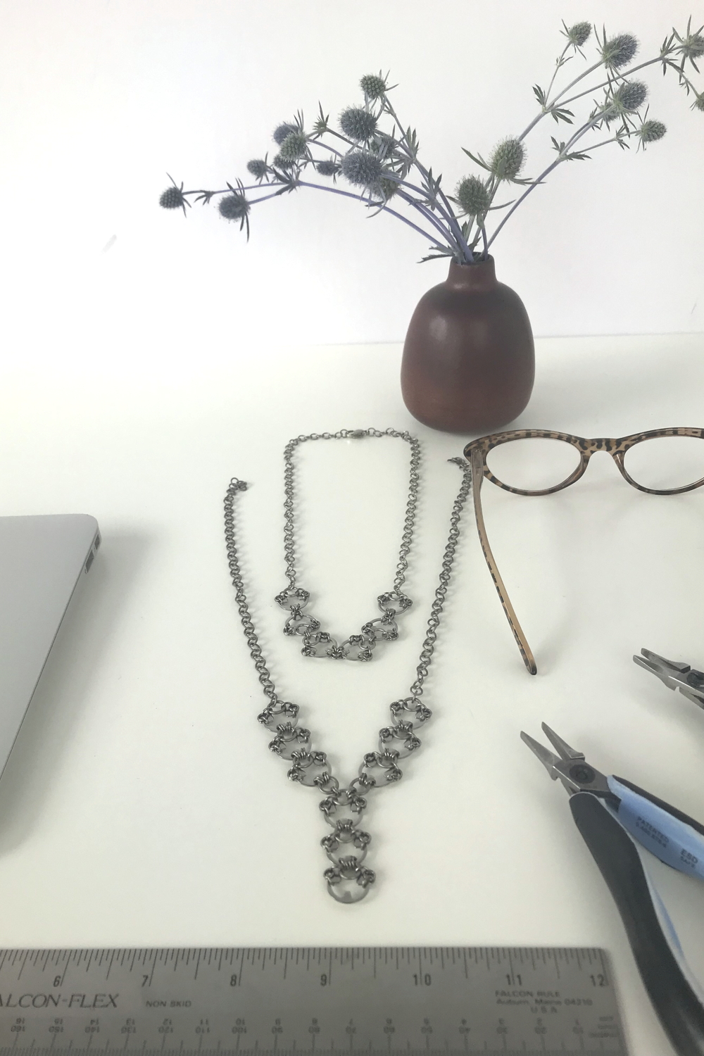 Sea holly (Eryngium) in a brown bud vase by Heath Ceramics on a desk next to Wraptillion's Delicate Scarab Necklace, Delicate Lotus Necklace, reading glasses, ruler, pliers, and a laptop.