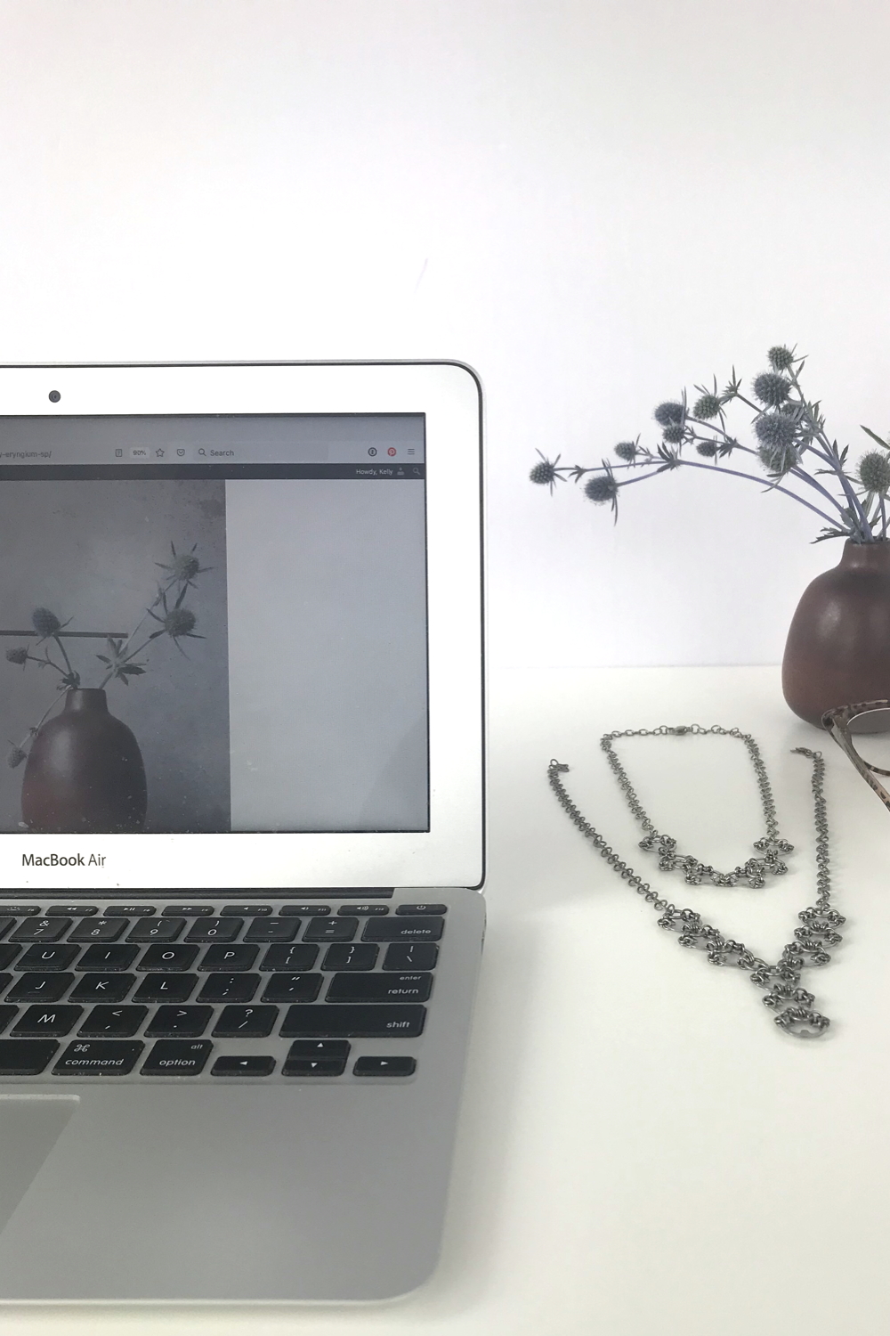 Sea holly (Eryngium) in a brown bud vase by Heath Ceramics on a desk next to Wraptillion's Delicate Scarab Necklace, Delicate Lotus Necklace, reading glasses, and a laptop.