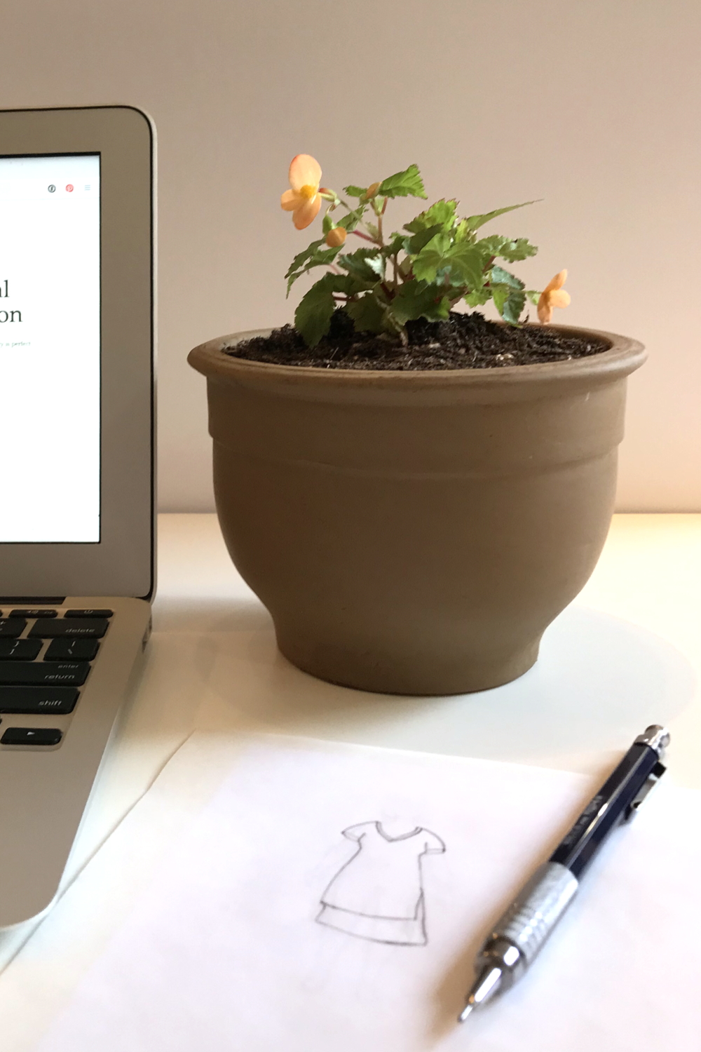 A small potted plant with orange flowers (Begonia sutherlandii) sits on a desk next to an open laptop, a pencil, and a fashion sketch of a dress.