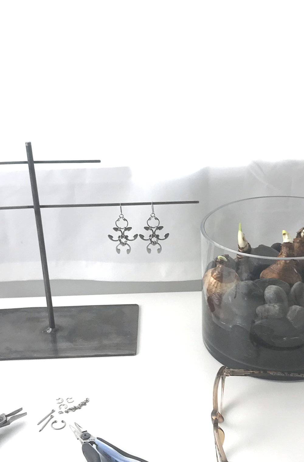 Studio vignette: a white desk in front of a white curtain, with a sneak peek of Wraptillion's jewelry piece in process with pliers, engineering components, and titanium jump rings, next to leopard reading glasses, a jewelry stand with Wraptillion's spiky abstract statement Garland Earrings and a cylindrical glass vase with black rocks and paperwhite narcissus bulbs just beginning to sprout