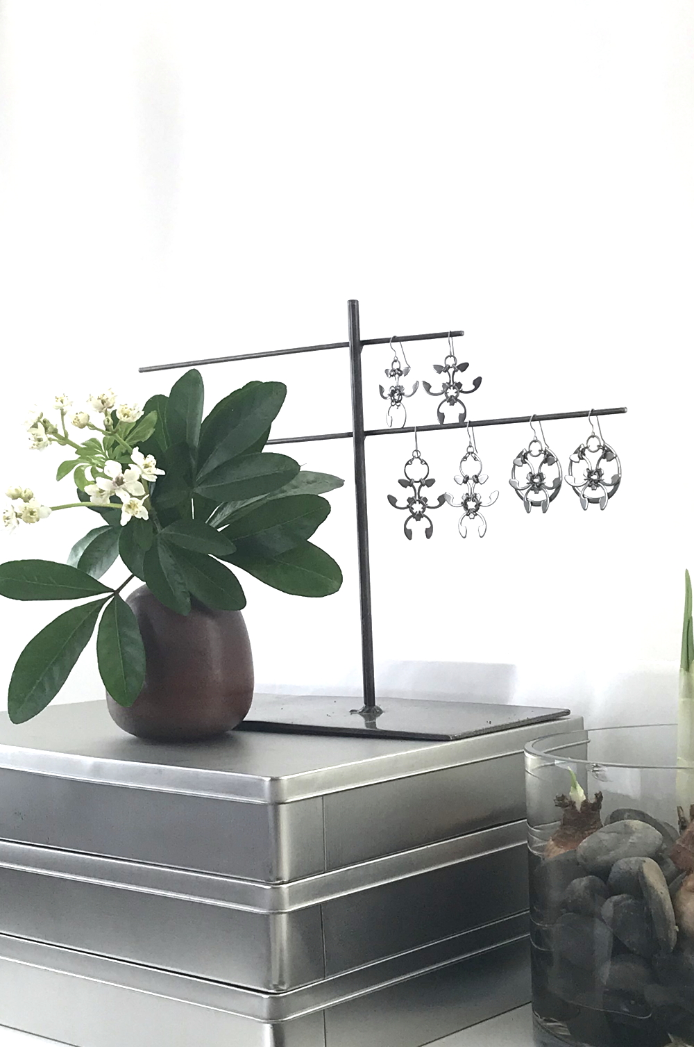 Studio vignette: a stack of metal boxes with a brown bud vase of Choisya ternata (Mexican orange blossom) green leaves and small white flowers, next to a welded steel jewelry stand with Wraptillion's modern abstract botanical statement earrings: the Trellis, Garland, and Rose Window styles. A glass cylinder vase with rocks and paperwhite narcissus bulbs sits nearby, in front of a white curtain.