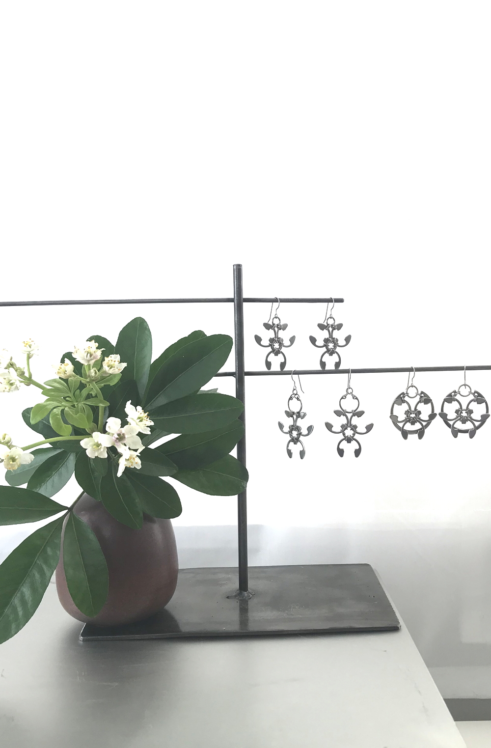 Studio vignette: a photo of a brown Heath Ceramics modern bud vase with choisya green leaves and white flowers, next to a welded steel jewelry stand with Wraptillion's spiky edgy Trellis Earrings, Garland Earrings, and Rose Window Earrings nearby, in front of a white curtain.