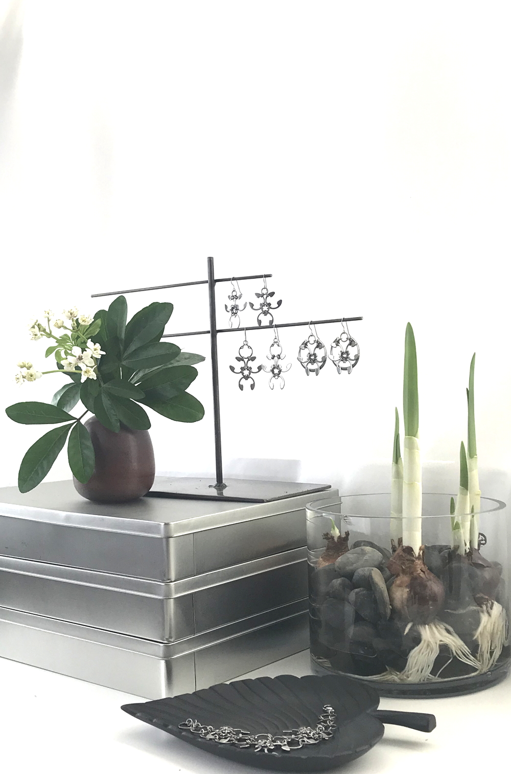 Studio vignette: a brown vase of Choisya ternate (Mexican Orange Blossom) flowers sits on a stack of silver-colored metal paper boxes next to a glass cylindrical vase of gray rocks and paperwhite bulbs (narcissus forced in water and stones), with Wraptillion's spiky abstract botanical statement Trellis Earrings, Garland Earrings, and Rose Window Earrings dangling from a jewelry stand nearby, and a black leaf-shaped dish holding the Garland Bracelet.