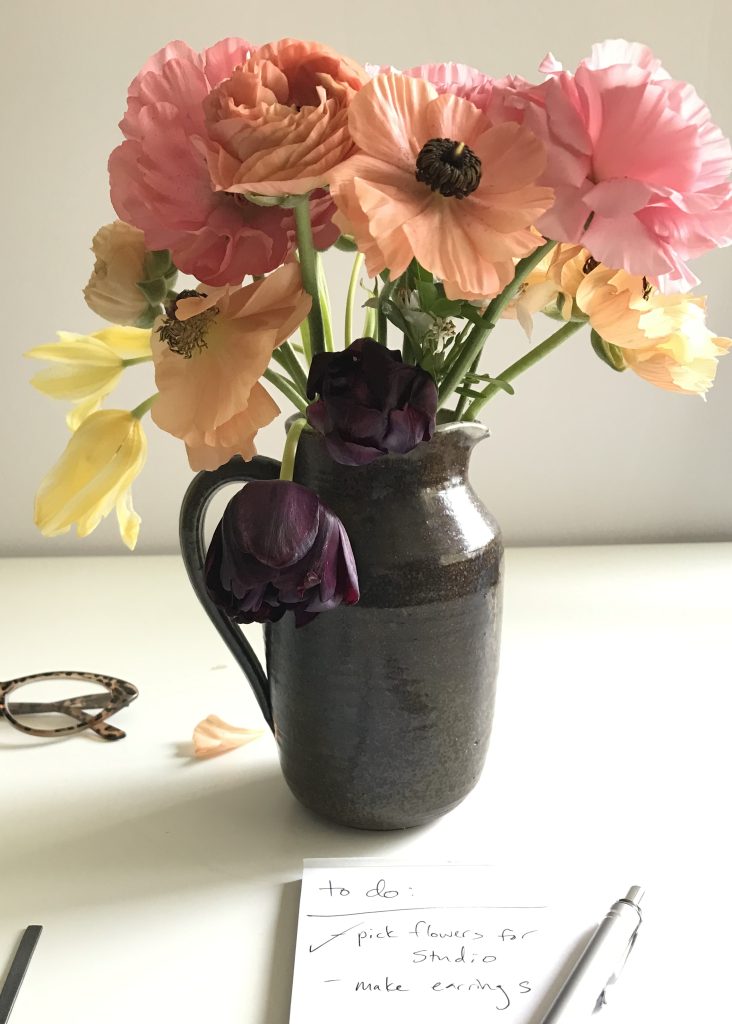 Salmon and pink ranunculus with drooping black and pale yellow tulips in a green ceramic pitcher, on a white desk with reading glasses and a to do list. The list reads: to do: pick flowers for studio; make earrings.