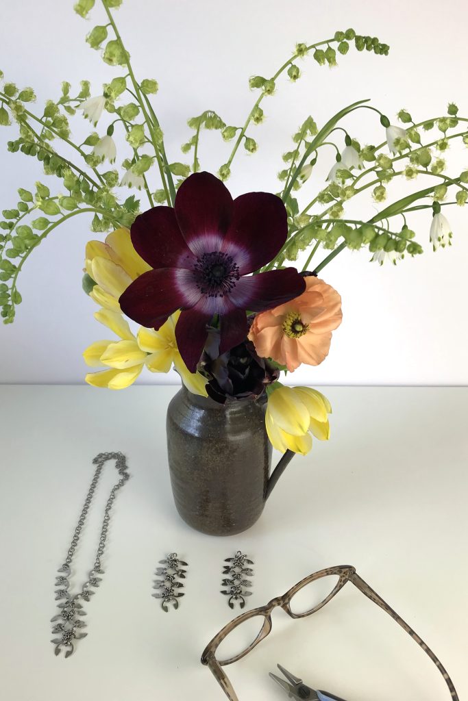 A bouquet of yellow and black tulips with salmon ranunculus, green tellima, white snowflakes, and a burgundy anemone in a hand-thrown glazed ceramic pitcher, on a white desk next to Wraptillion jewelry in progress, with glasses and tools.