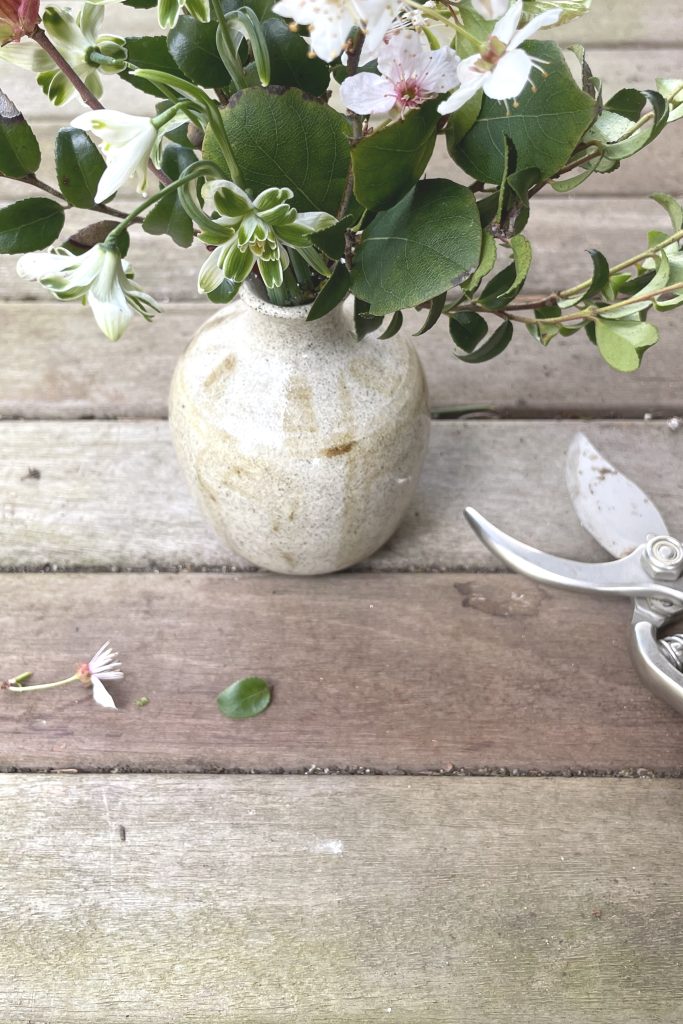 A small arrangement of late winter flowers in a handmade ceramic vase, on a worn wooden surface, next to pruners.