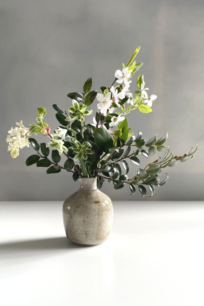 A small arrangement of late winter flowers on a white desk, including double snowdrops, plum flowers, and evergreen huckleberry.