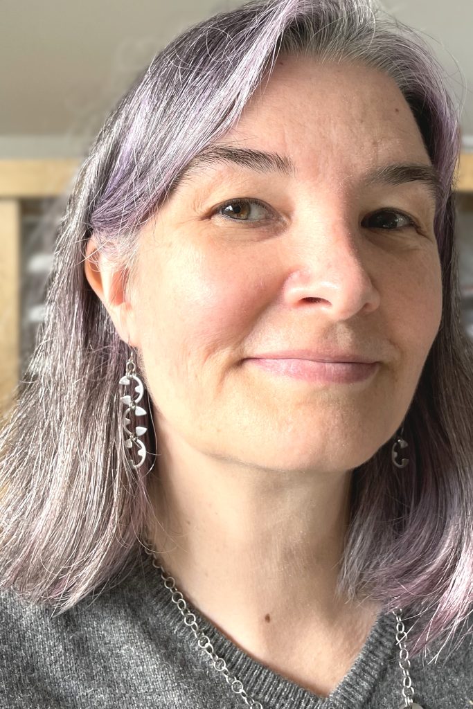 A white woman with purple hair and modern silver-colored earrings.